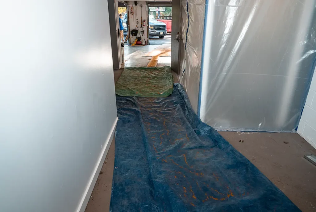 Narrow hallway with blue and green tarps on the floor, leading to a garage with tools and a truck outside. Plastic sheets cover the right wall.
